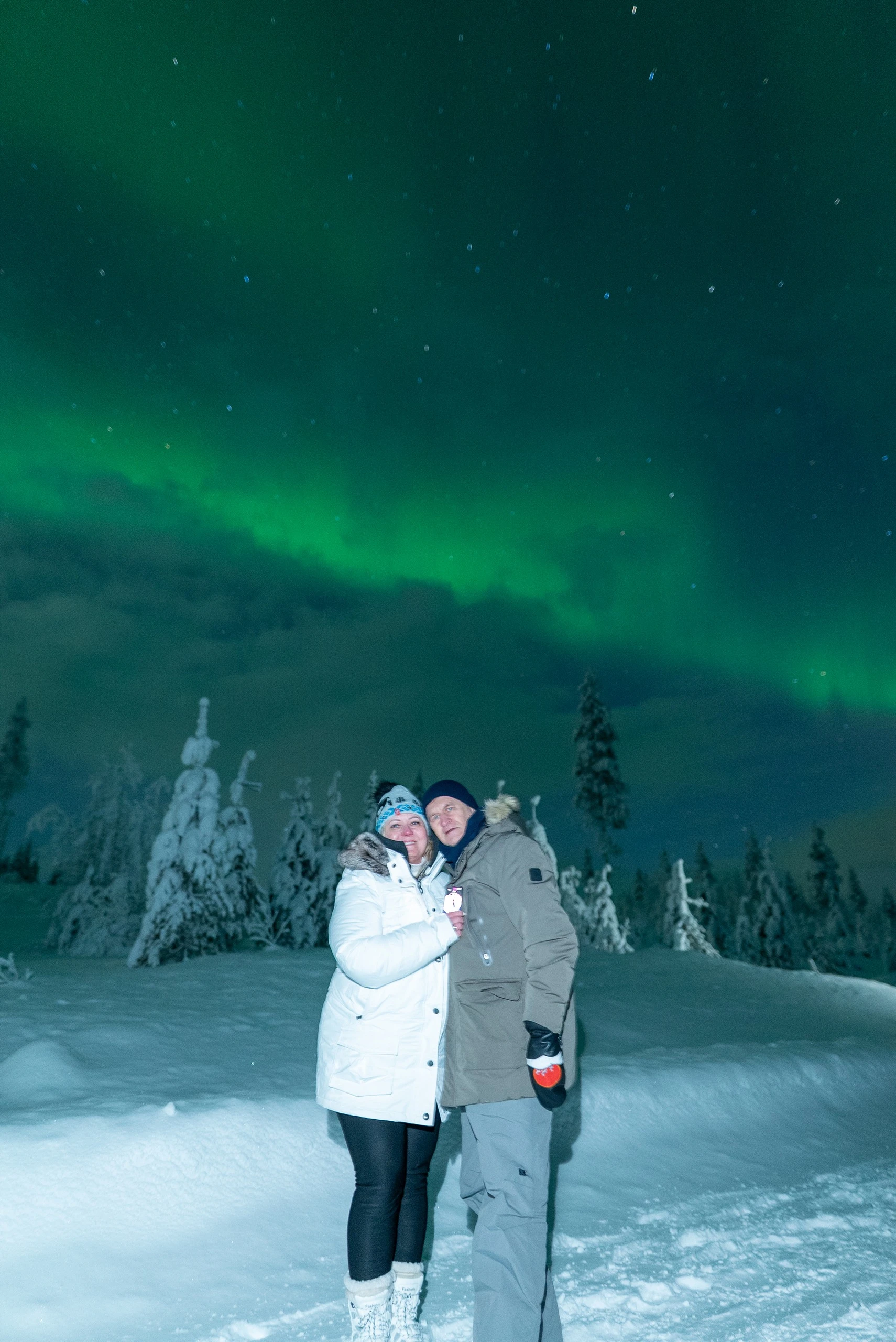 Couple in front of snow-crowned trees under the aurora borealis